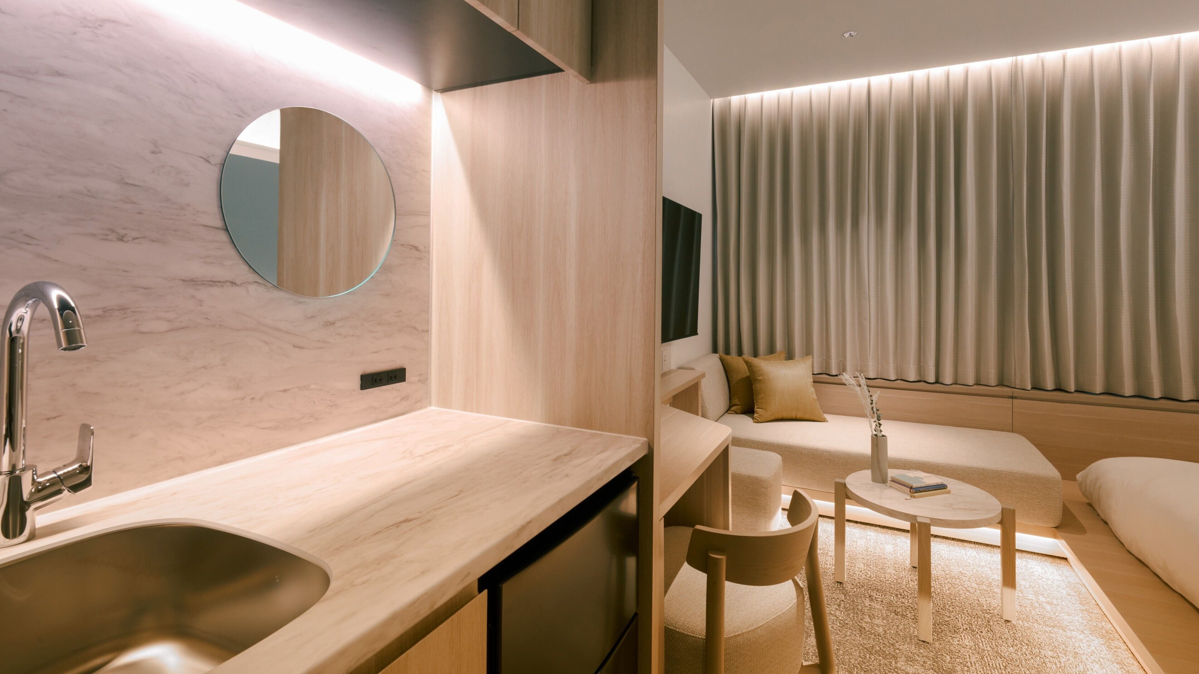 A modern hotel room with a marble sink, wooden counter, round mirror, and a seating area with a sofa and coffee table.