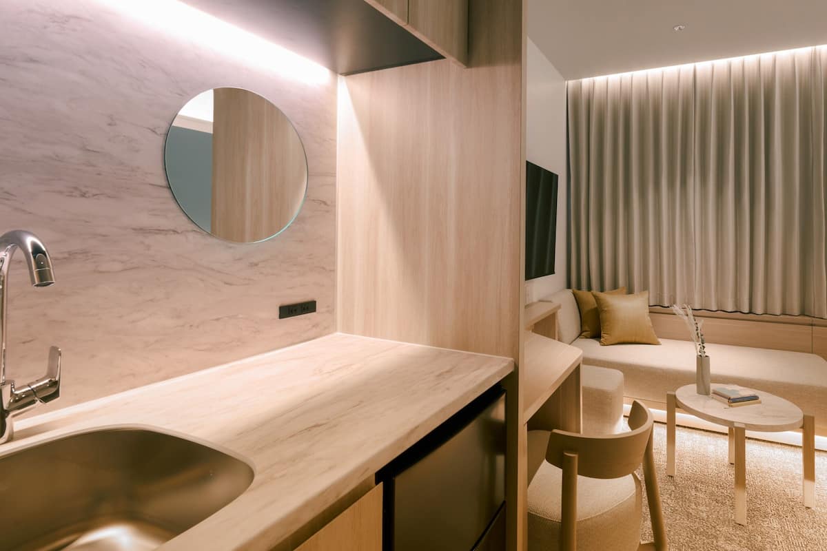 A modern hotel room with a marble sink, wooden counter, round mirror, and a seating area with a sofa and coffee table.