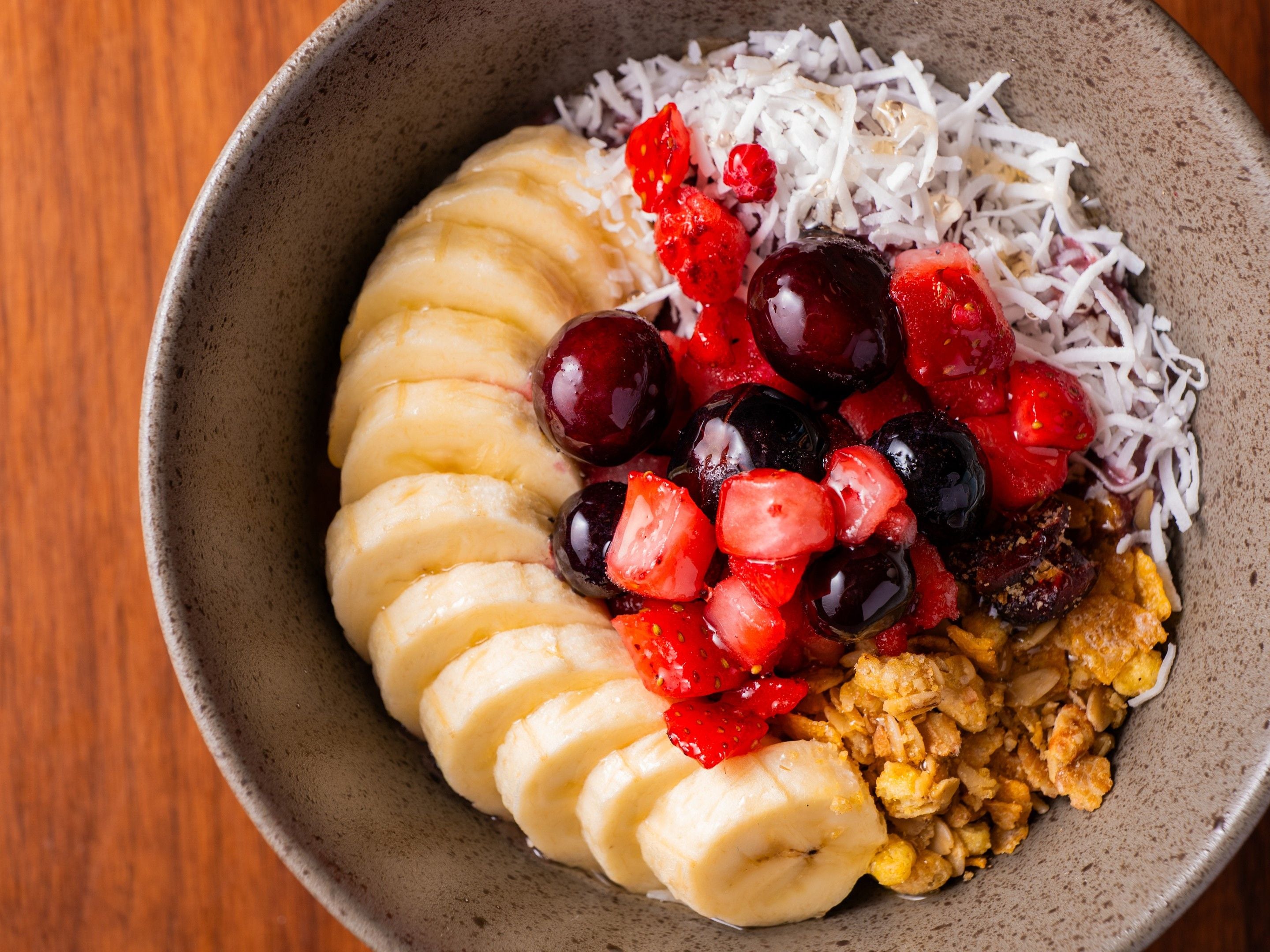 A bowl containing sliced bananas, red and black grapes, shredded coconut, and granola.