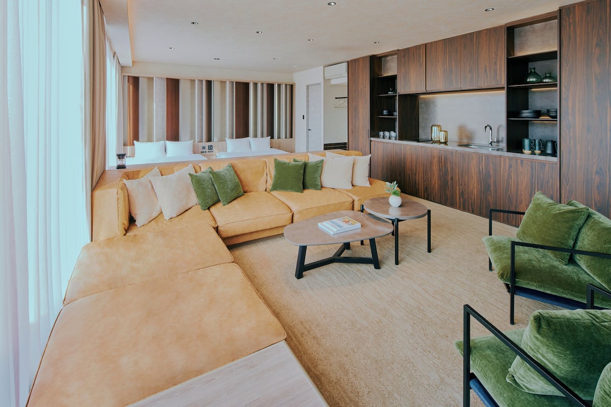 A modern living room with a large sofa, a coffee table, and a green armchair. There is a kitchen area with wooden cabinets and a sink.