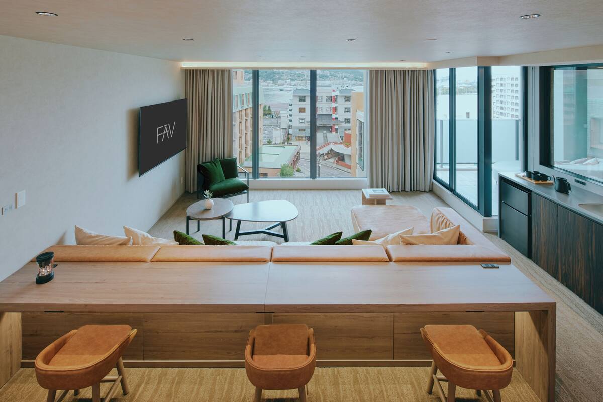 A modern hotel room with a large sofa, a small table, and a view of the city through large windows.