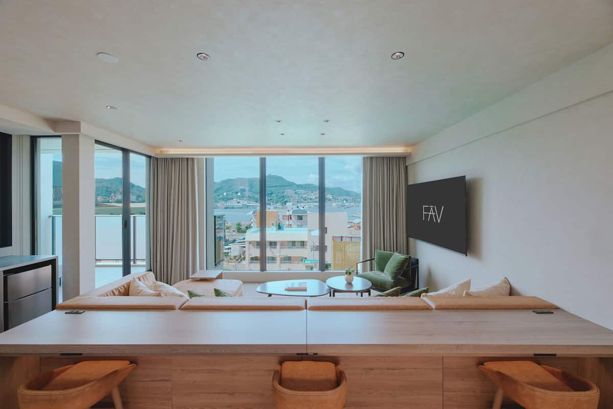 A modern hotel room with a large window, a sofa, a small table, and a TV mounted on the wall.