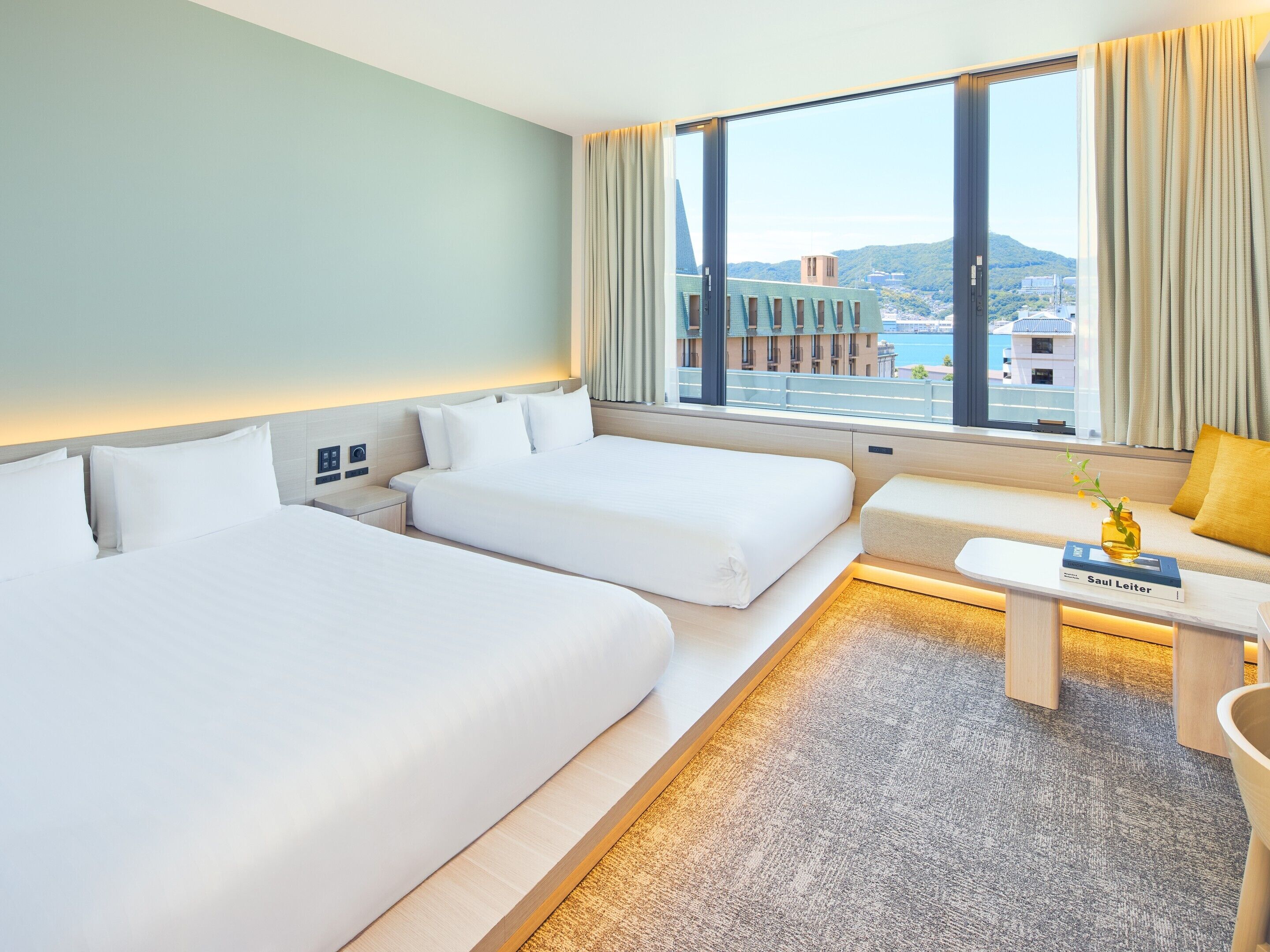 Habitación de hotel con dos camas, un sofá, una pequeña mesa con un libro y vistas a edificios y al agua.
