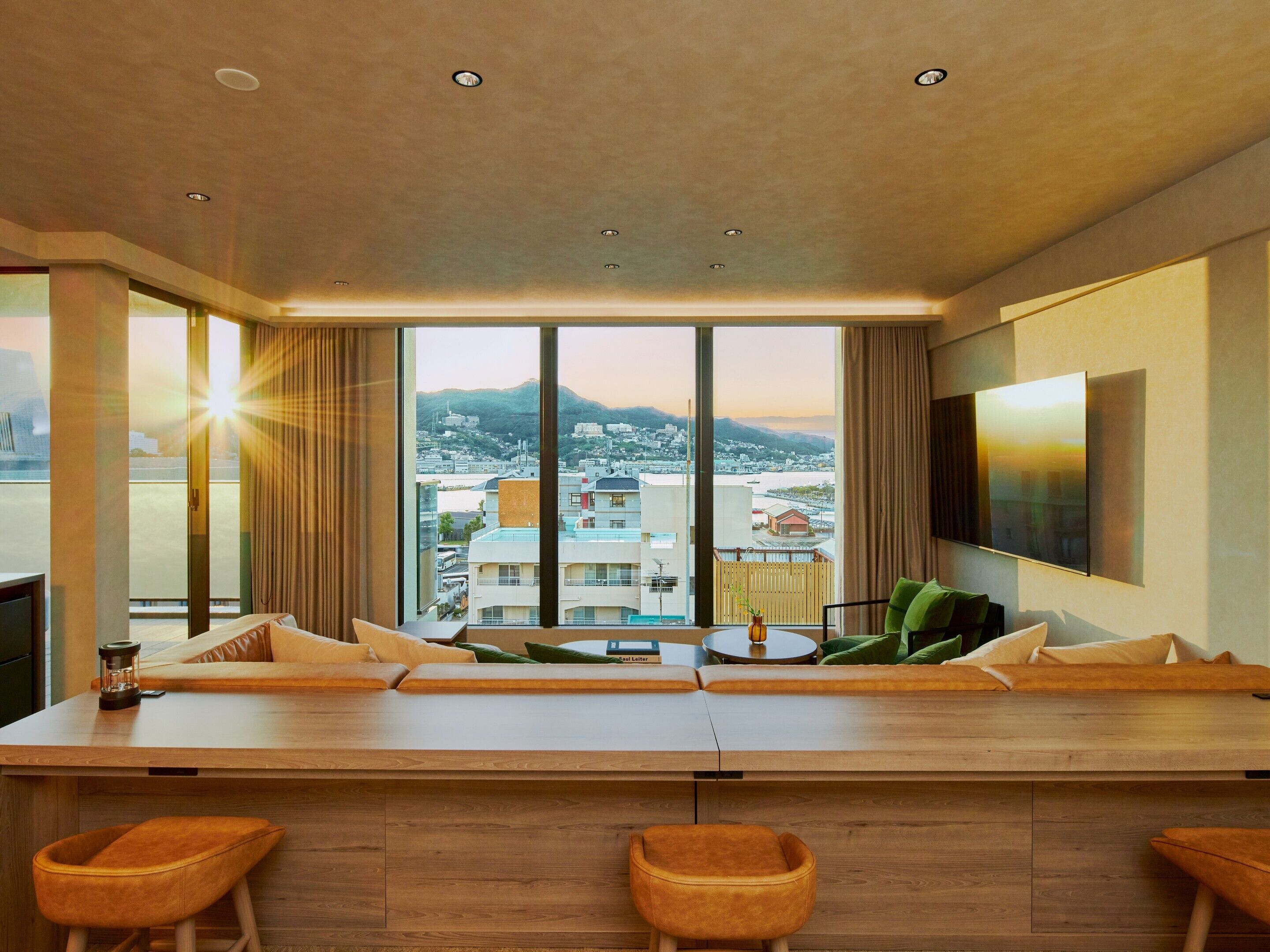 A modern hotel room with a large window, a flat-screen TV, a wooden bar area, and a city view.