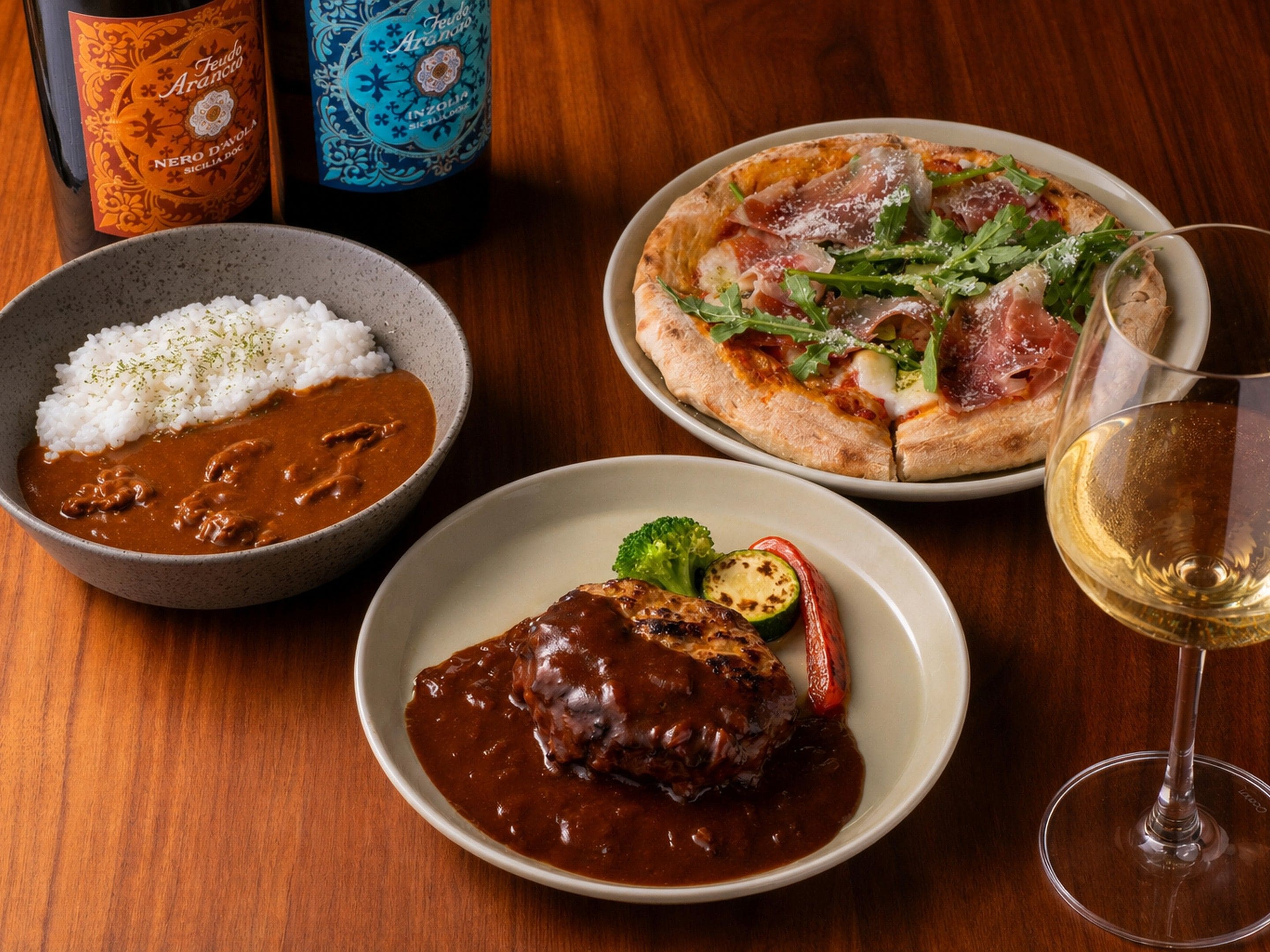 A table with a variety of dishes including rice with curry, a pizza with vegetables and meat, and a steak with sauce. There are also two bottles of wine and a glass of wine on the table.