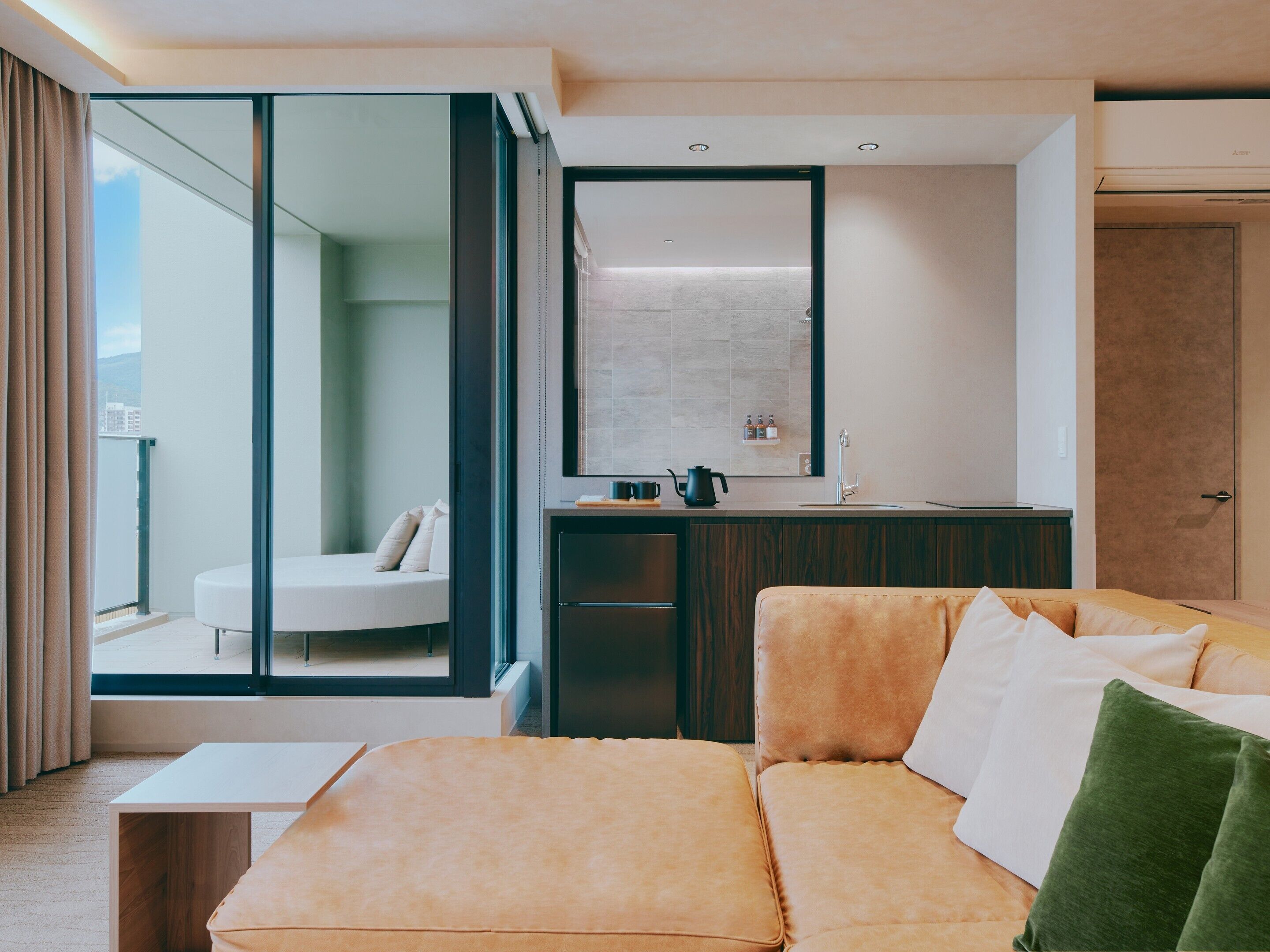 A modern hotel room with a sofa, a small table, a mirror, and a view of the city.