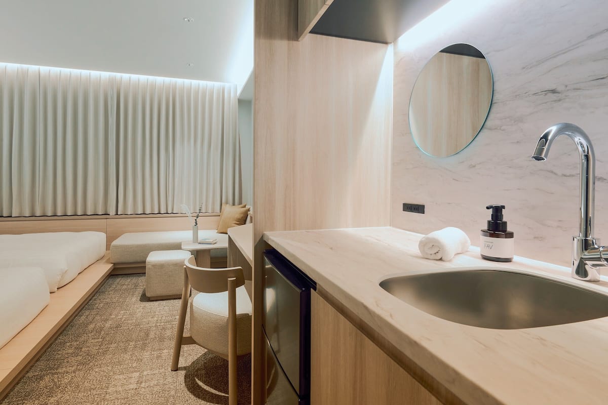 A modern hotel room with a bed, a seating area, a bathroom with a sink and mirror, and a window with curtains.