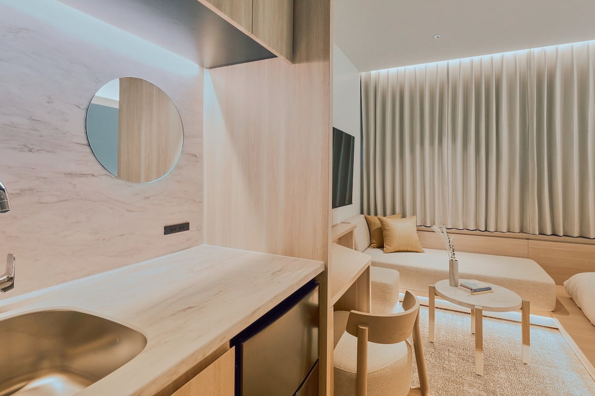 A modern hotel room with a wooden vanity, a round mirror, a sofa, a small round table, and a large window with curtains.