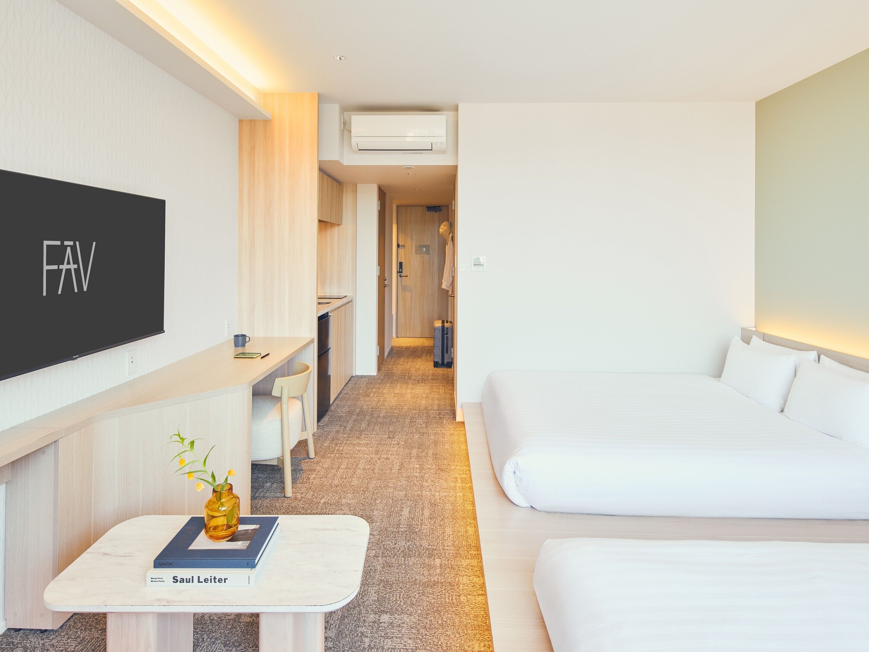 A hotel room with a bed, a desk with a chair, a TV, and a small table with a book and a vase.