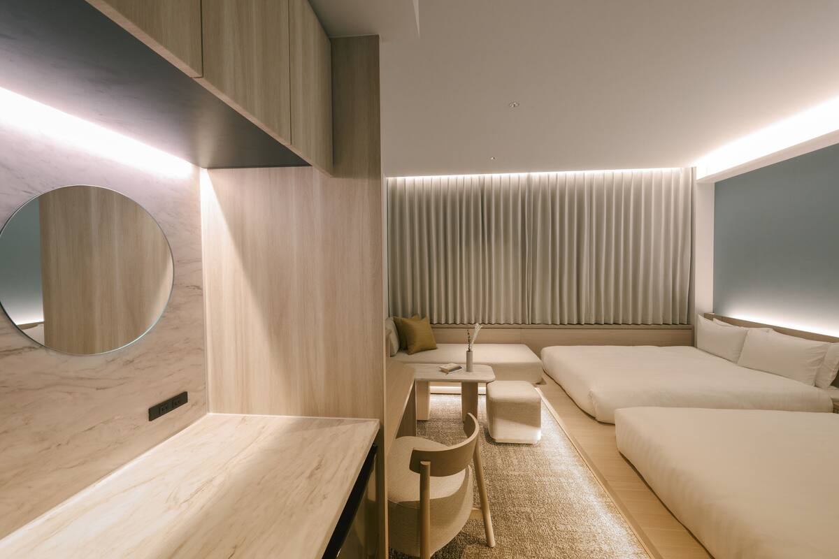 A modern hotel room with a wooden floor, a round mirror, a sofa, a small table, and a large window with curtains.