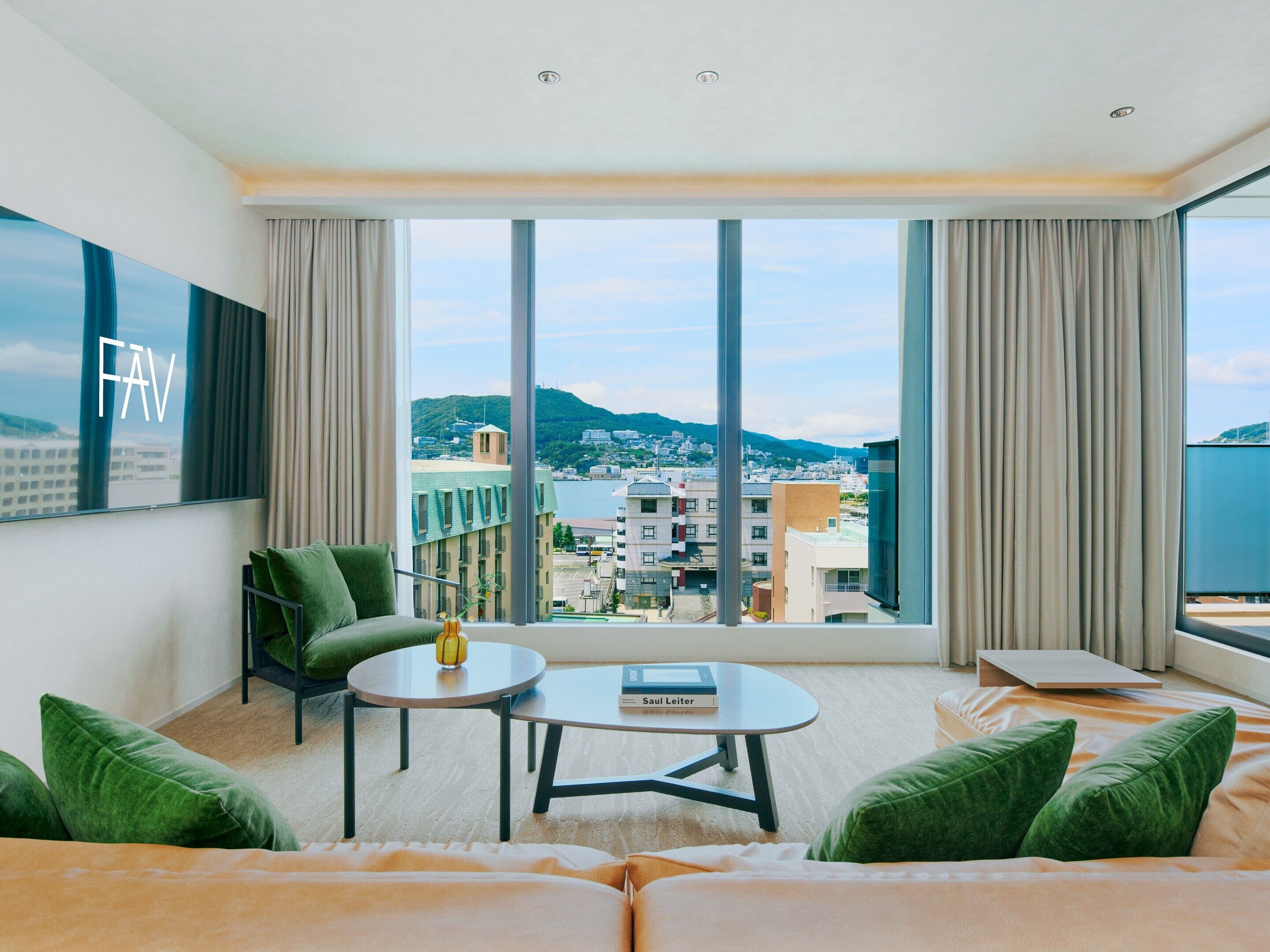A modern hotel room with a large window, a sofa, a small table, and a green chair.