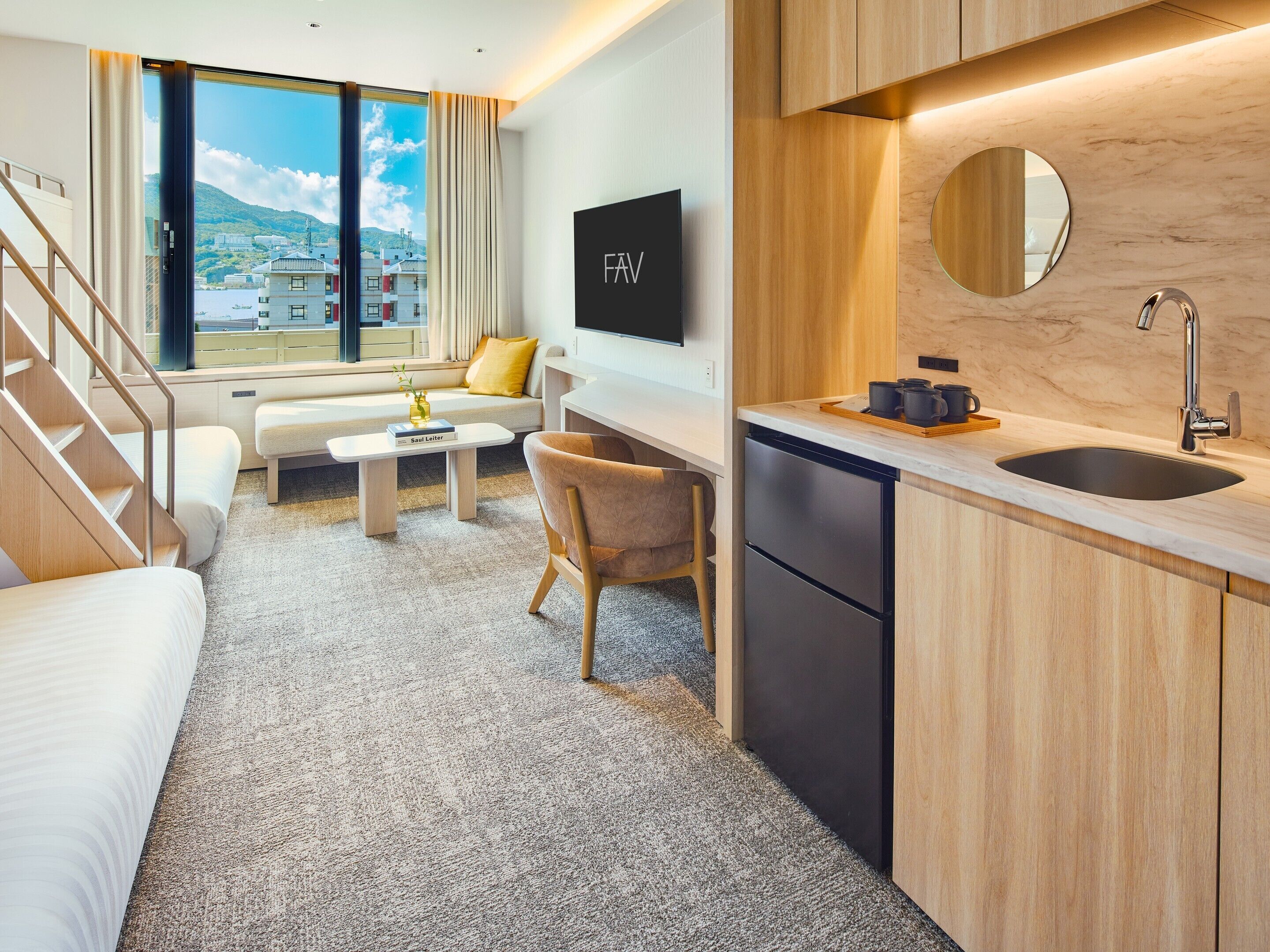 A modern hotel room with a sofa, a small table, a chair, a kitchen area with a sink, and a large window offering a view of buildings and hills.