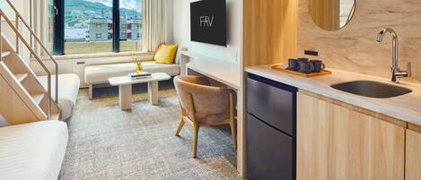 A modern hotel room with a sofa, a small table, a chair, a kitchen area with a sink, and a large window offering a view of buildings and hills.