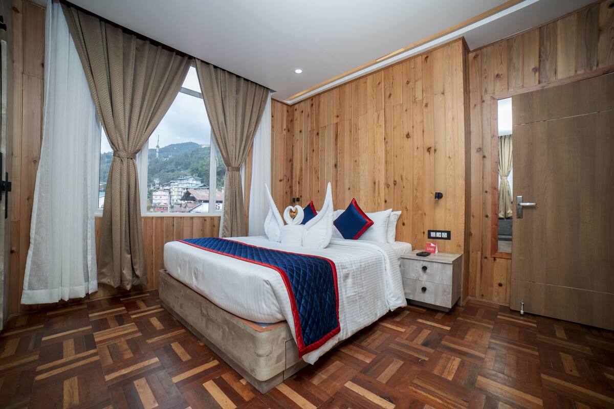 A hotel room with a large bed, wooden panel walls, a nightstand, and a view of a cityscape.
