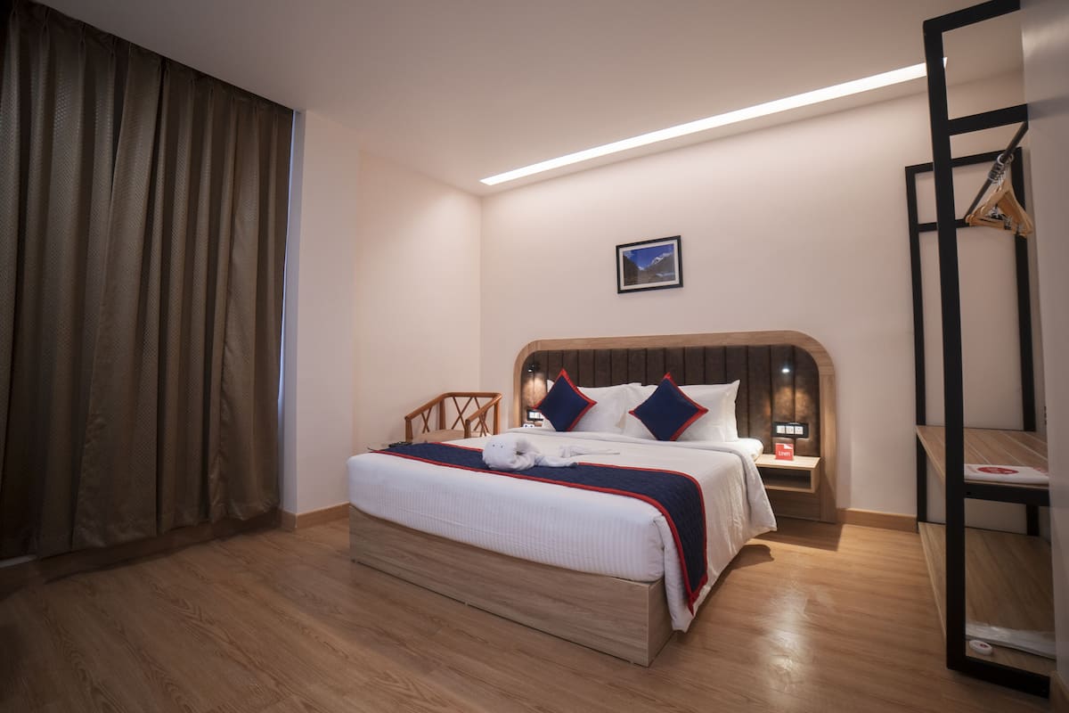 A neatly arranged hotel room with a wooden floor, a bed with a white and navy blue bedspread, a nightstand with a lamp, and a wall-mounted shelf.