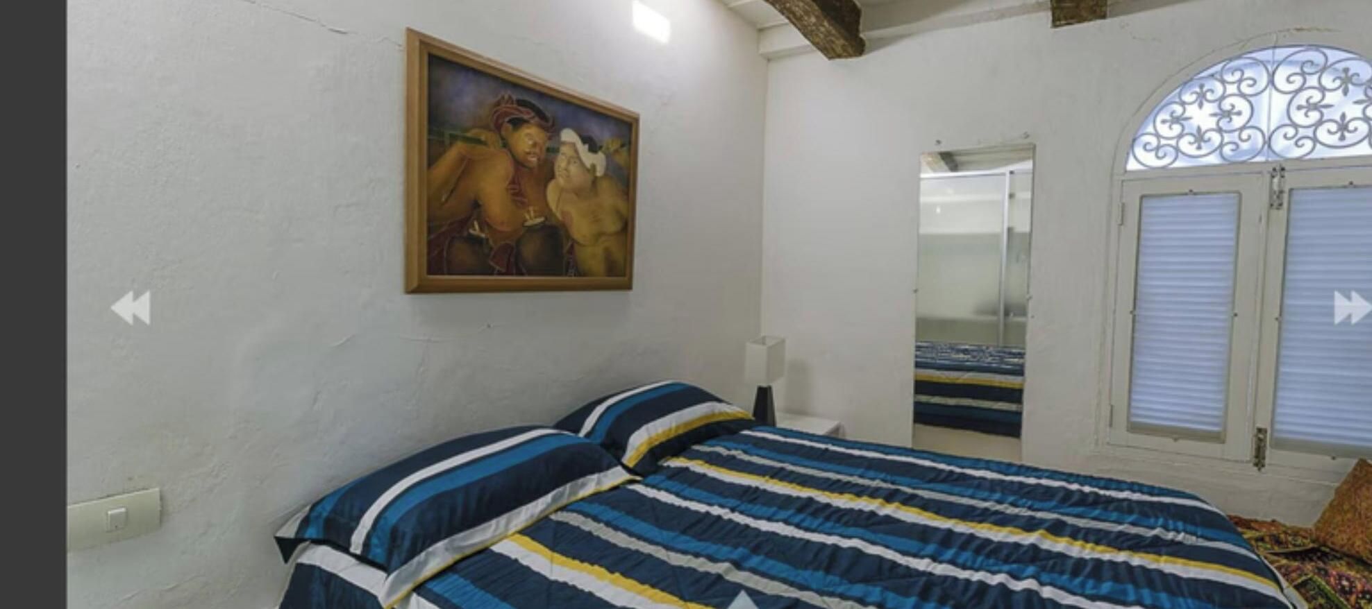 Ein Schlafzimmer mit einem Bett, einem Gemälde, einem Fenster mit Jalousien und einem Spiegel.