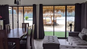 A room with a dining table, chairs, a sofa, and a fan. There is a view of a beach and a thatched-roof structure outside. - Crystal Cove (Sipalay)