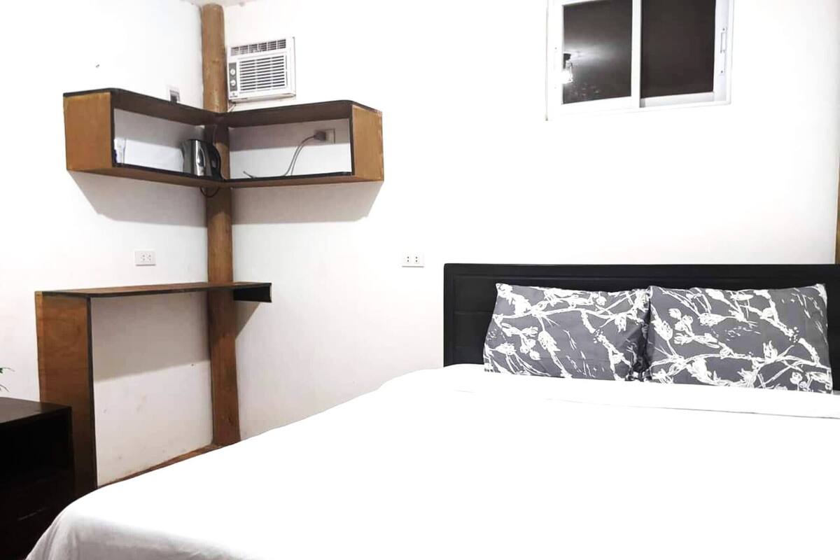 Un dormitorio con cama, mesita de noche, estante en la pared y ventana.