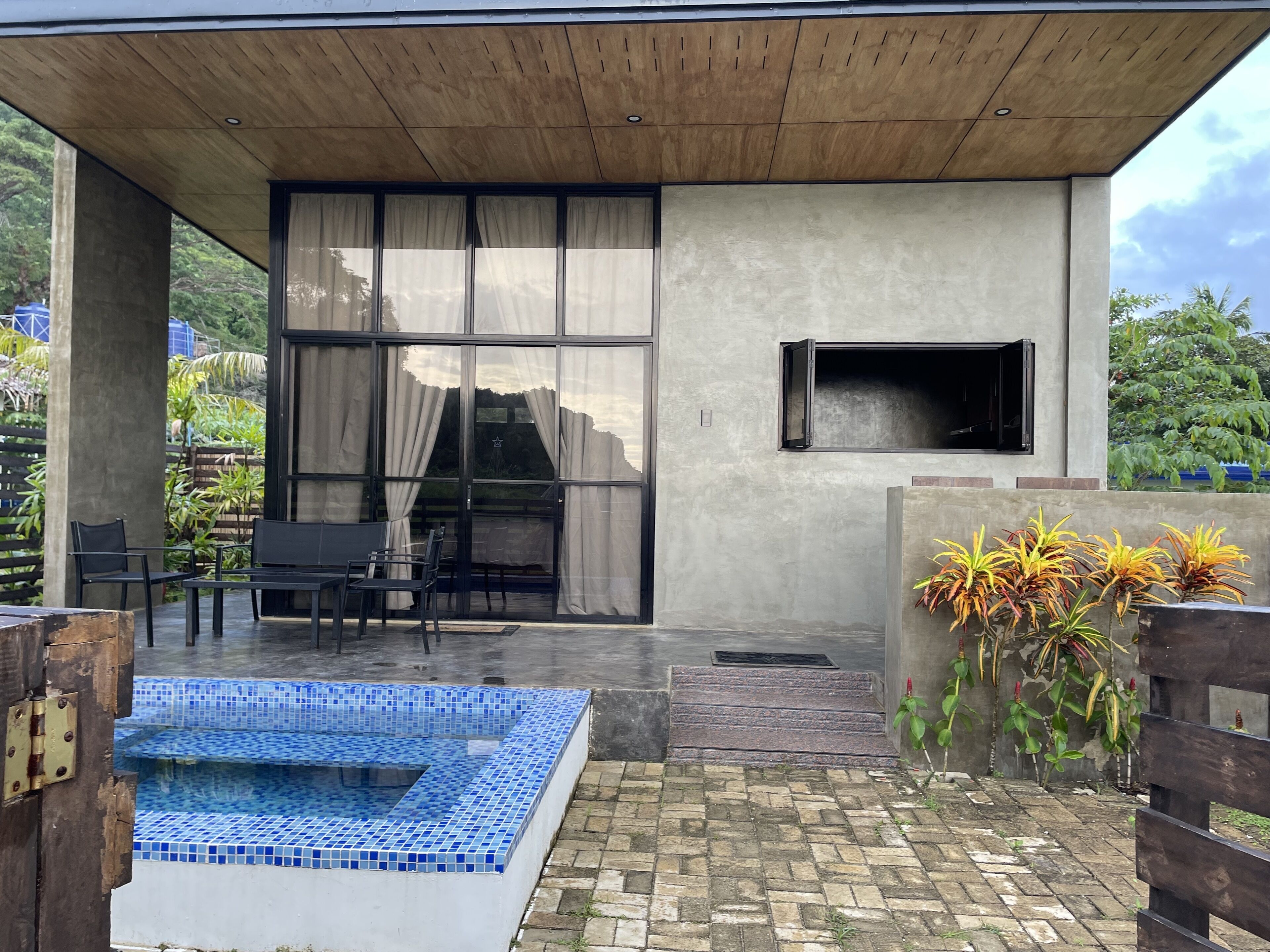 A modern outdoor pool area with a tiled blue pool, black outdoor furniture, and a concrete wall with a window and a mounted TV.