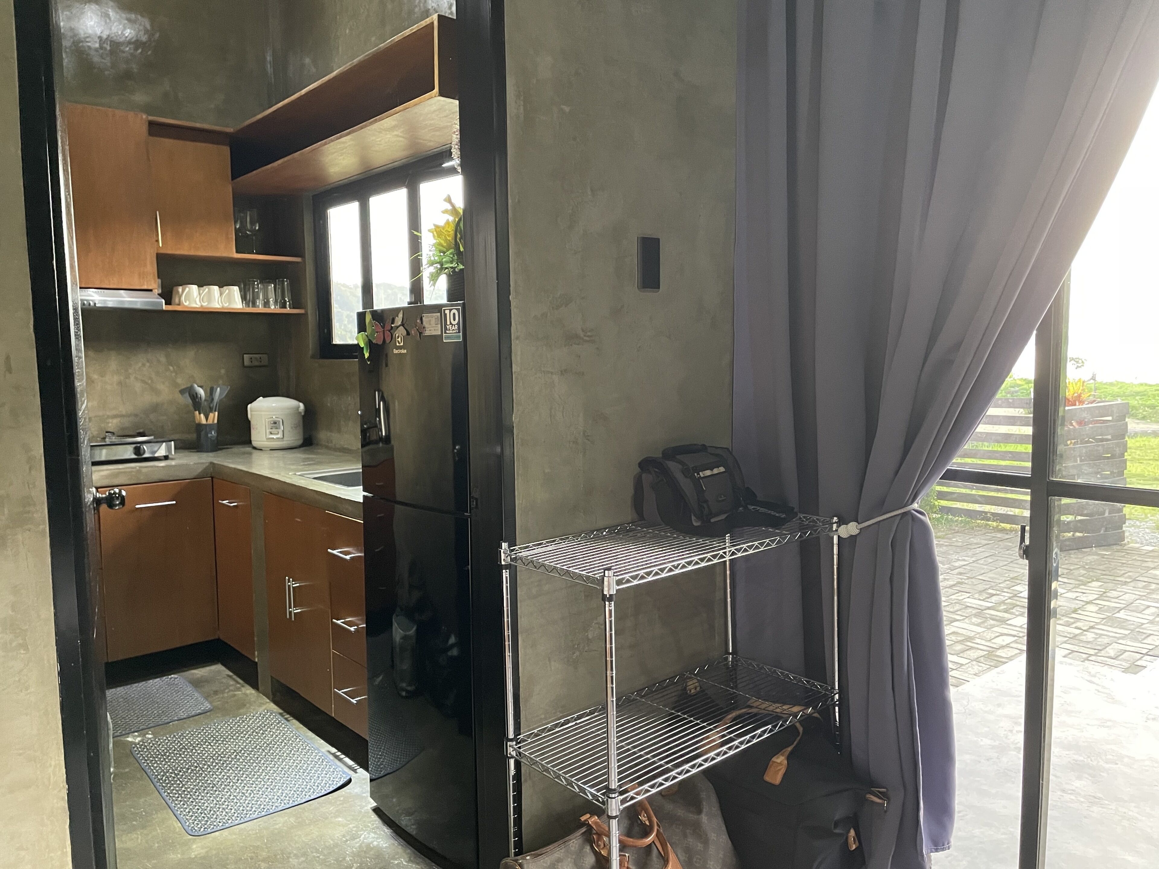 A compact kitchen with wooden cabinets, a black refrigerator, and a small countertop. There is a metal shelf unit against the wall and a window with curtains.