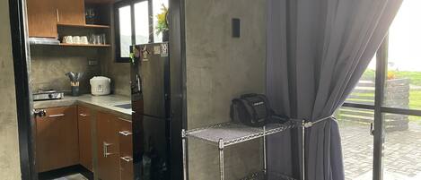 A compact kitchen with wooden cabinets, a black refrigerator, and a small countertop. There is a metal shelf unit against the wall and a window with curtains.