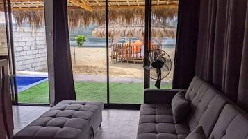 A room with a grey sofa, a fan, and a view of a pool and outdoor area with a thatched roof.