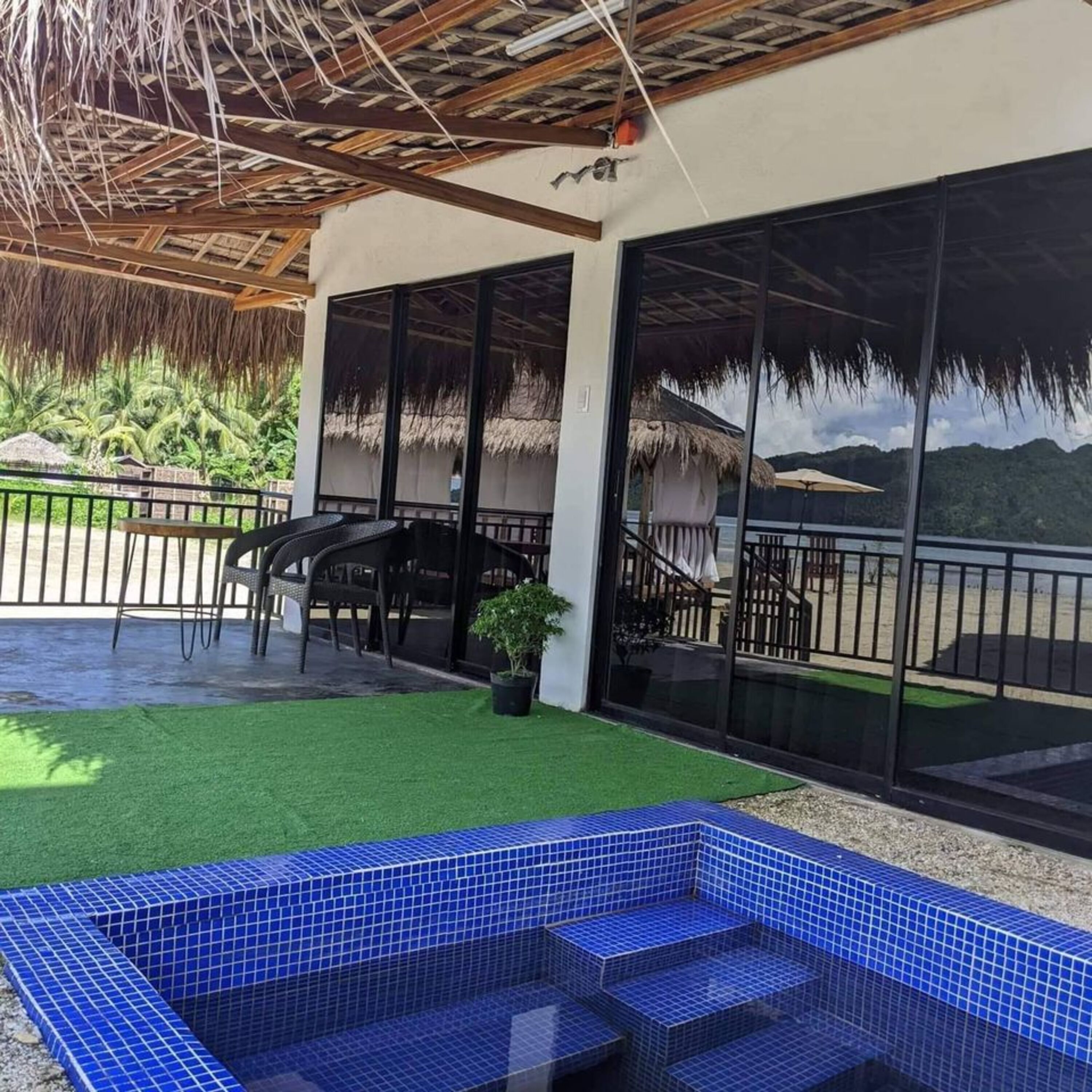 Un espace piscine avec des carreaux bleus, un coin salon couvert avec des chaises, et une vue sur une chaîne de montagnes à travers de grandes portes vitrées.