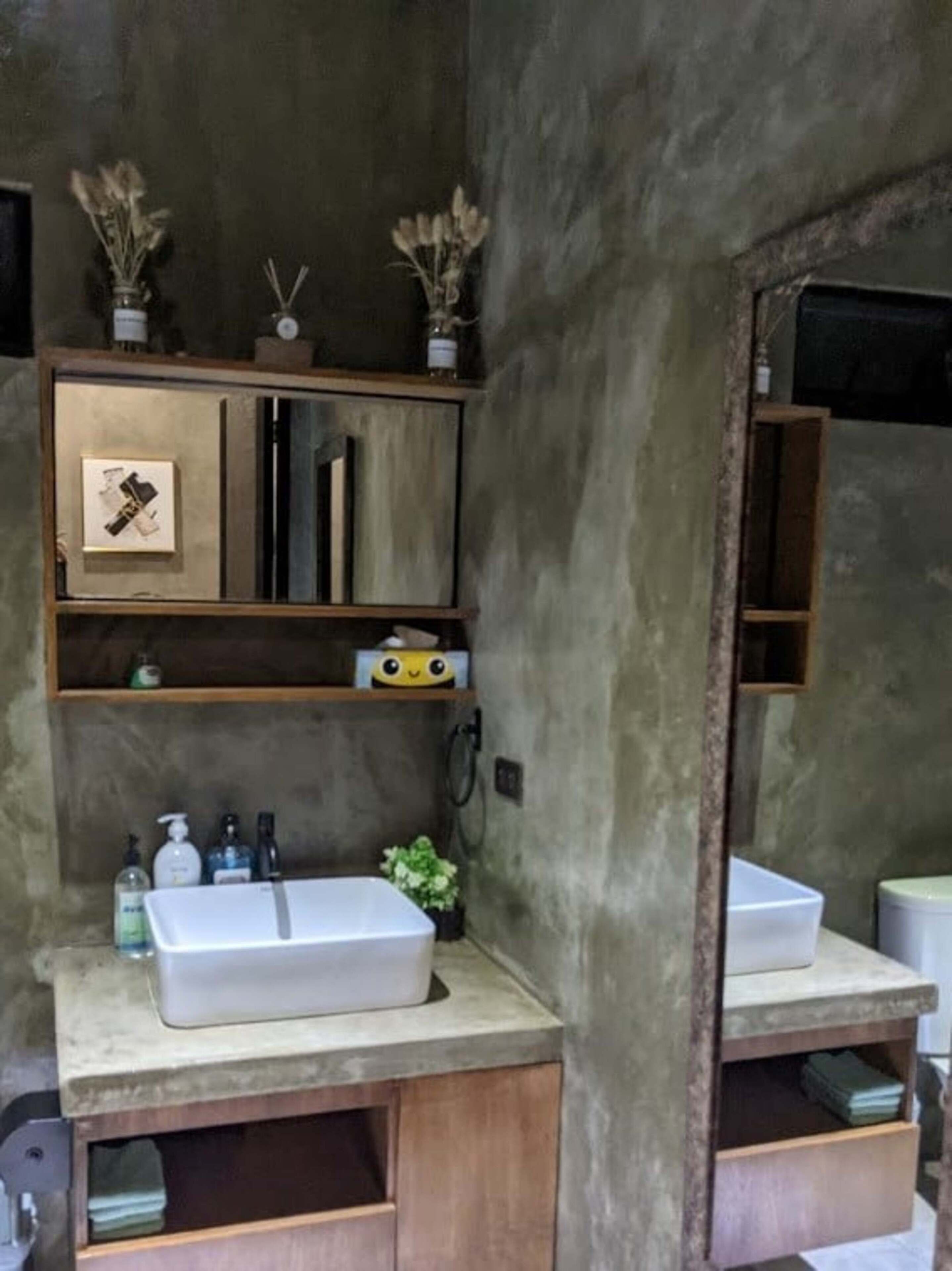 Casa de banho com um espelho grande, uma pia branca e um toucador de madeira. Existem duas pequenas plantas em vasos sobre o toucador e uma moldura com uma imagem no espelho.
