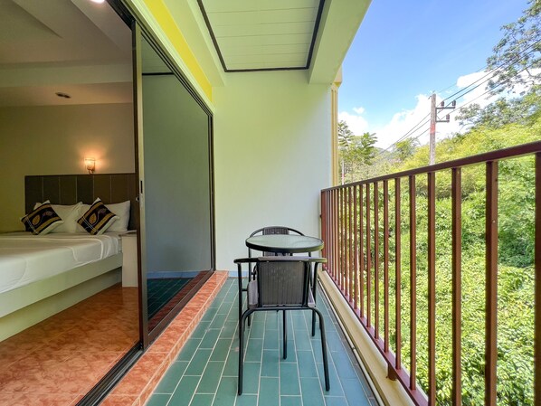 A balcony with a table and chairs, a bed with pillows, and a view of greenery. - Tri's Miracle Kata Beach Side (Karon)