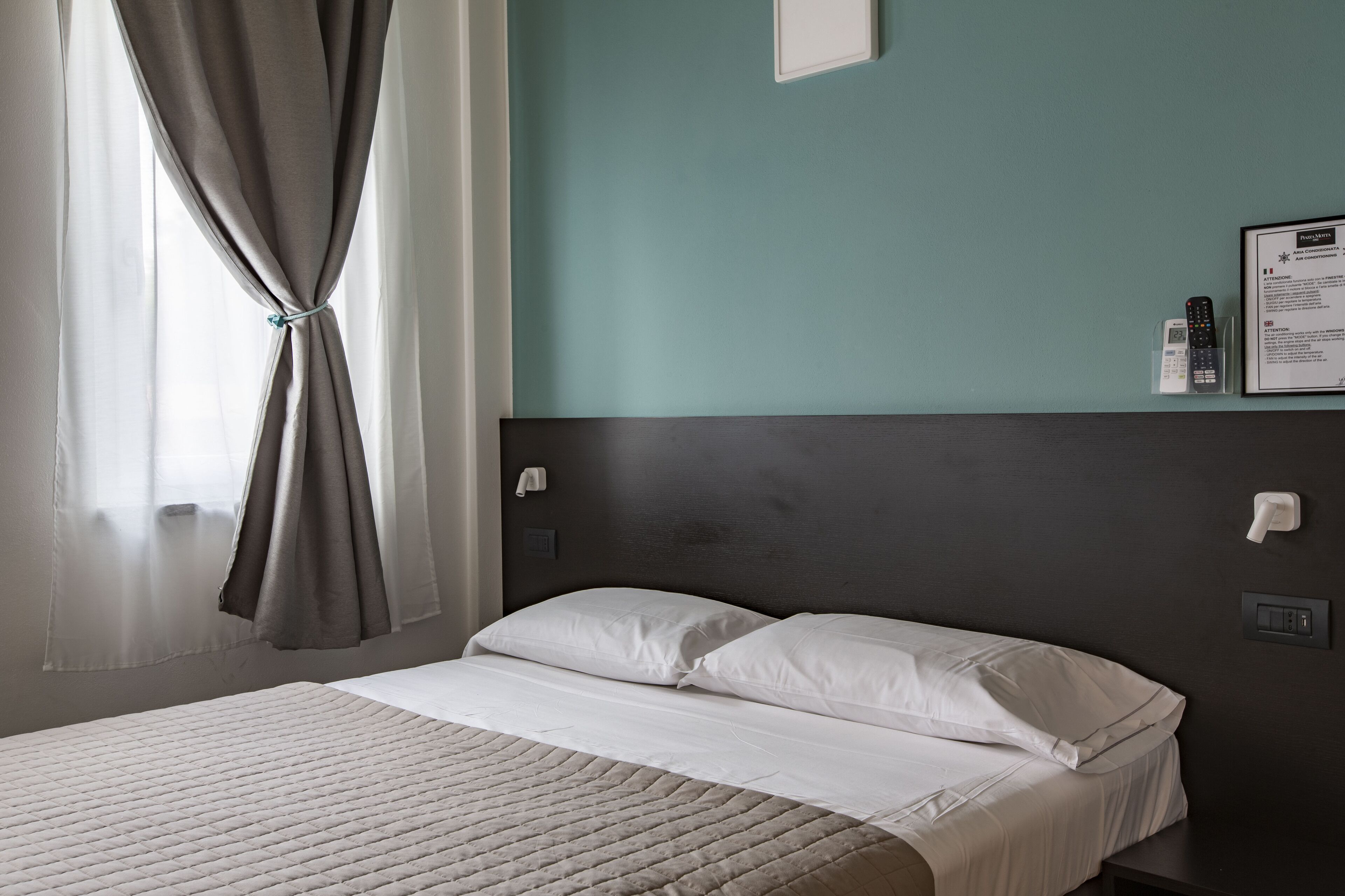 A hotel room with a bed, curtains, a wall-mounted phone, and a framed notice.