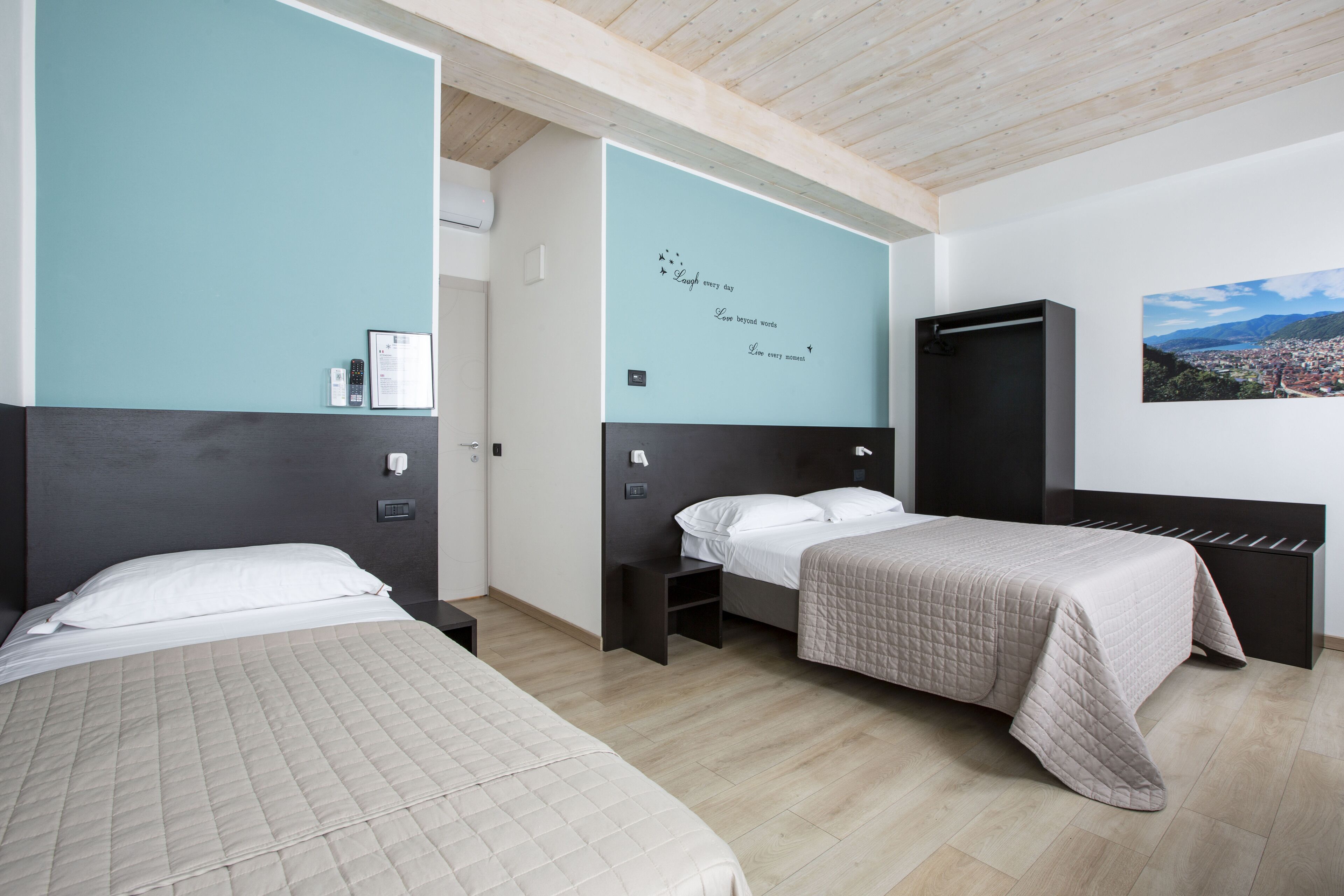 Une chambre d’hôtel avec deux lits, une armoire noire, un mur orné d’un tableau représentant un paysage urbain, et un mur d’accent de couleur turquoise sur lequel sont inscrites des citations inspirantes.
