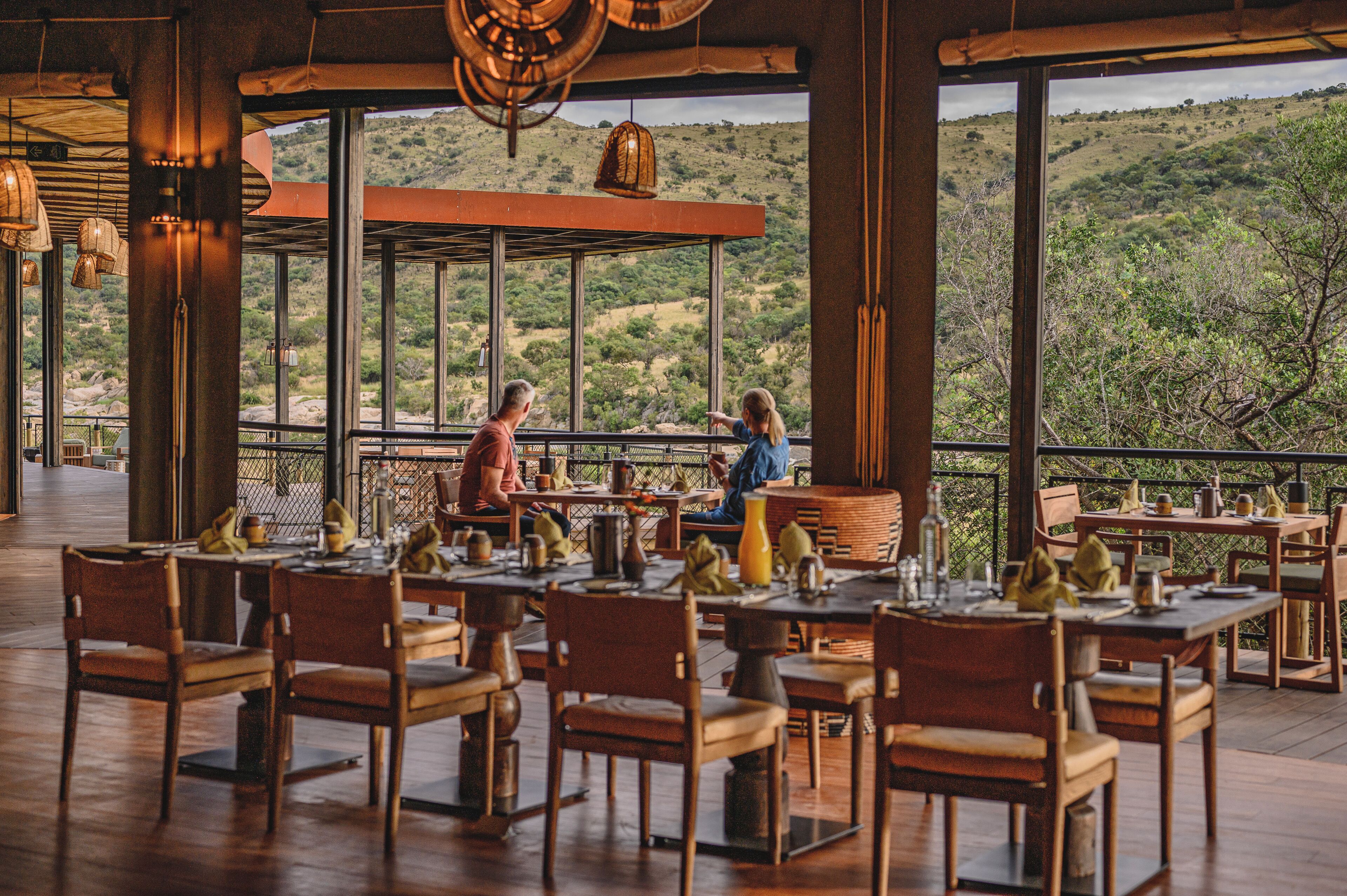 An outdoor dining area with tables and chairs, large windows offering a view of a hilly landscape.