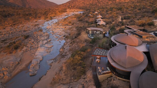 Un moderno complejo turístico con arquitectura singular, que incluye un edificio circular y varias estructuras en forma de cúpula, ubicado a orillas de un río en un paisaje desértico.