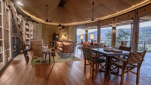 Un cuarto acogedor y bien iluminado, con piso de madera, una mesa de comedor con sillas, un sofá y una estantería. Amplias ventanas ofrecen vistas al exterior.