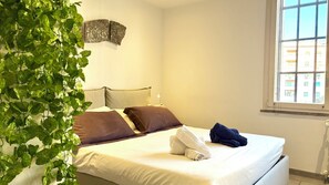 A bedroom with a bed, pillows, a bedside lamp, a window, and a plant.