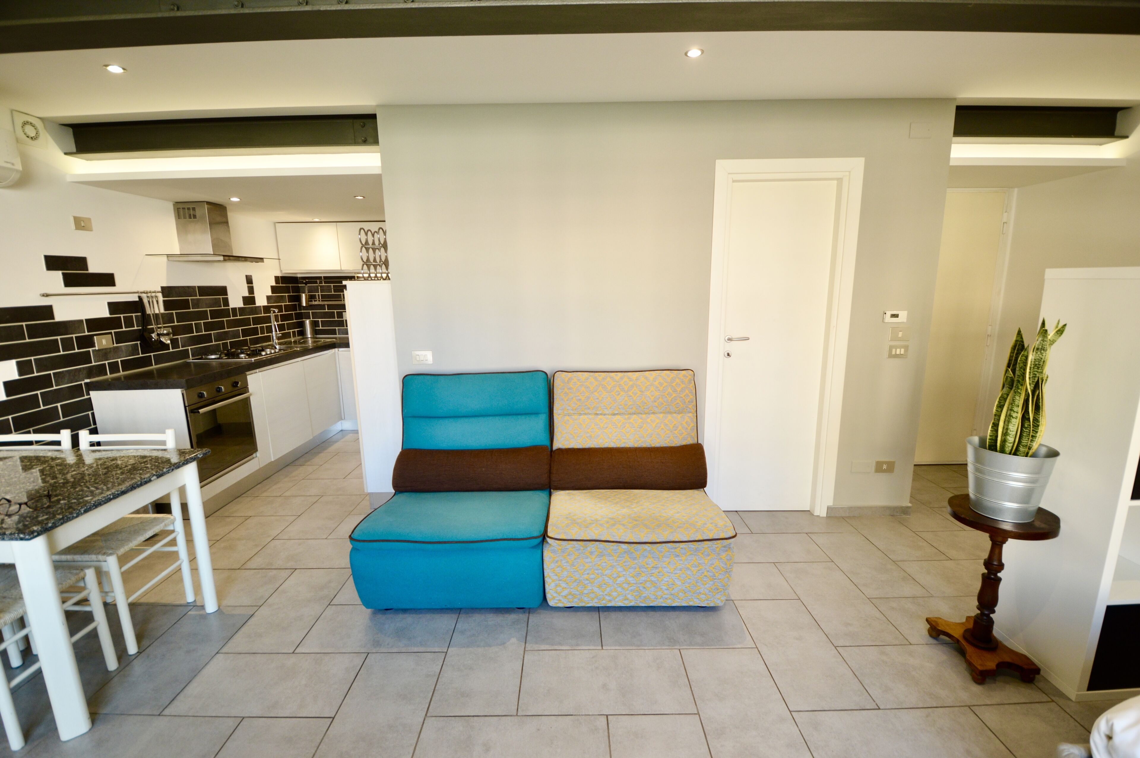 A modern kitchen with black and white tiles, a dining area with a white table and chairs, and a sofa with blue and beige cushions.