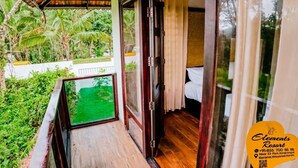 A balcony with a view of a green garden and palm trees, a wooden floor, and a bed with white bedding. - Elements Resort (Mananthavady)