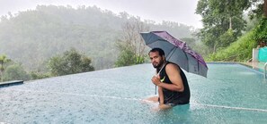 A person sitting on the edge of a pool with a purple umbrella, surrounded by a lush, forested hillside. - Elements Resort (Mananthavady)