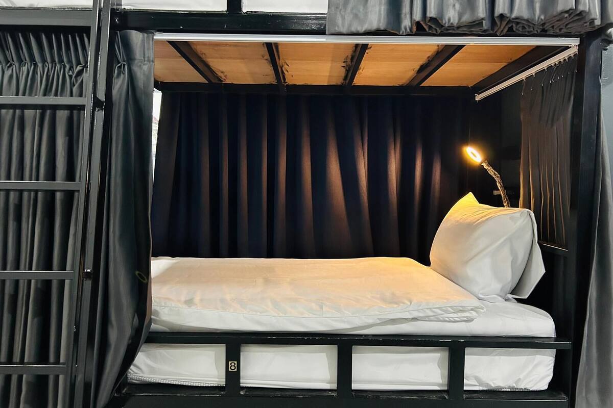 Une pièce de taille modeste, aux couleurs sombres, comprenant un lit simple, une petite table de chevet avec une lampe et une fenêtre recouverte d’un rideau.
