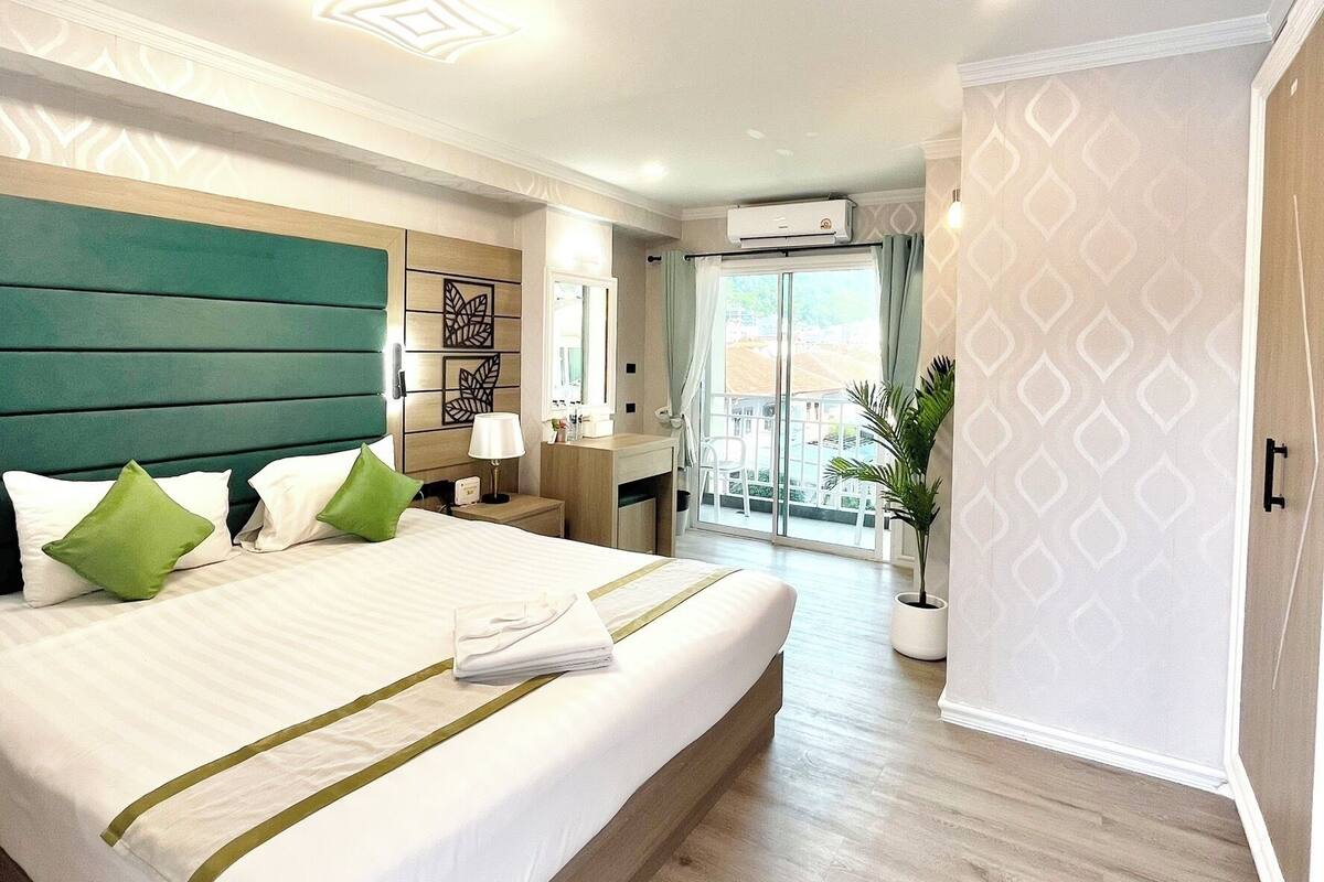 Une chambre d’hôtel moderne dotée d’un grand lit, d’une tête de lit en bois, d’un bureau avec une chaise, d’une plante en pot et offrant une vue sur l’extérieur grâce à une porte coulissante.