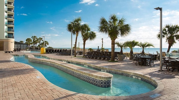 A pool area with a curved design, surrounded by lounge chairs and palm trees, with a building and ocean view in the background.