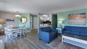 A modern living room with a blue sofa, white dining set, and a kitchen in the background.