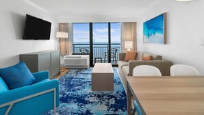 A hotel room with a balcony, a flat-screen TV, a sofa, a dining table, and a painting.