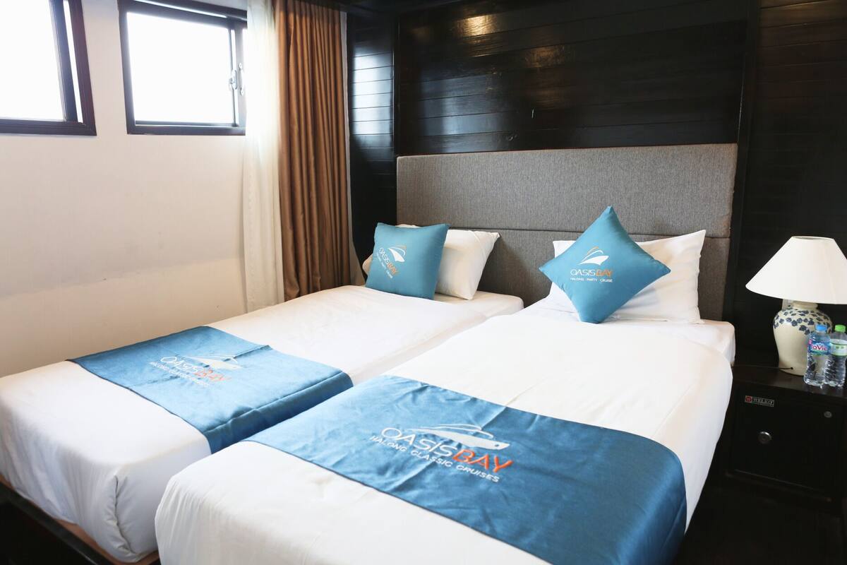 A hotel room with two beds, each with a blue Oasis Bay branded bedspread. There is a nightstand with a lamp and water bottles on it.