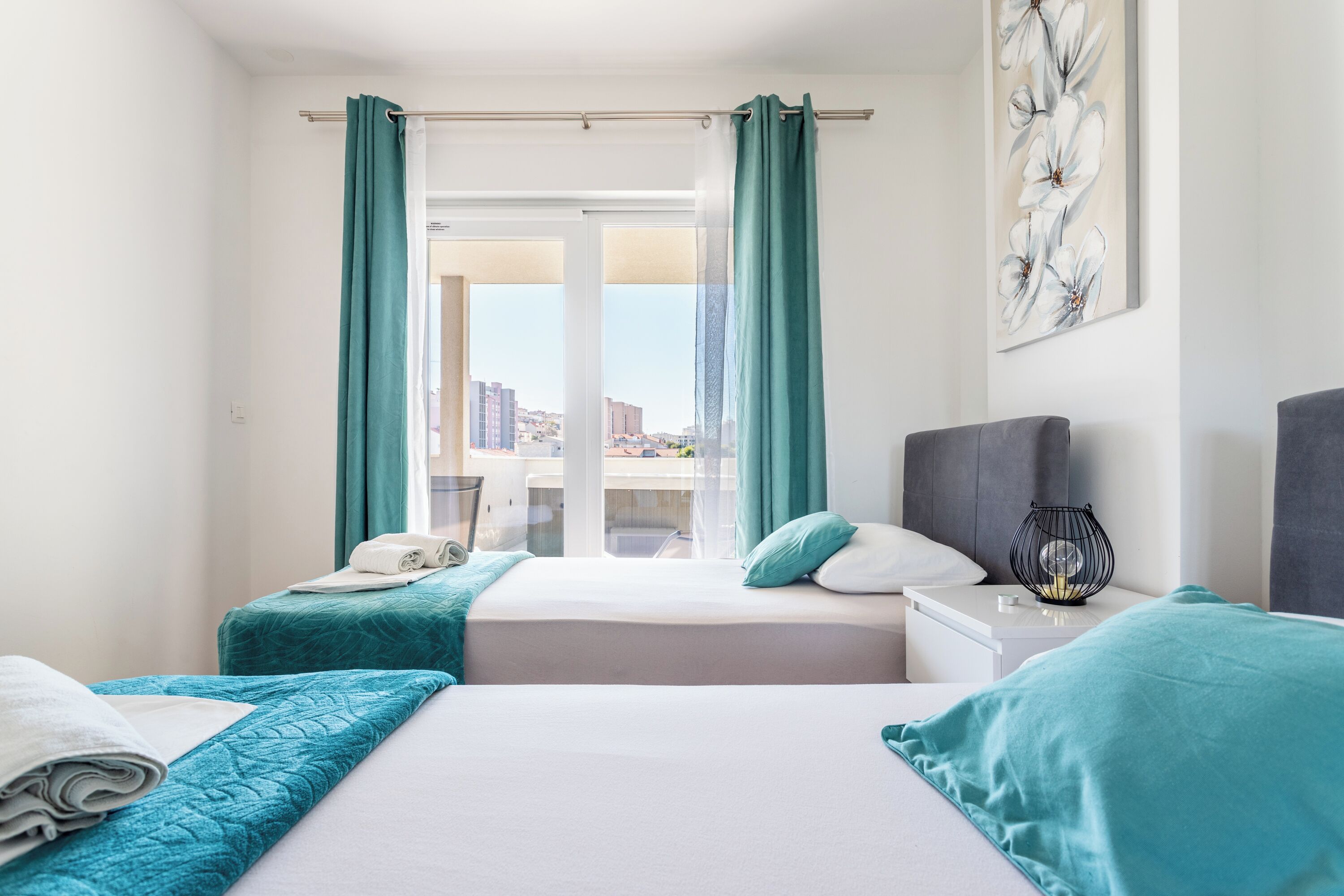 Habitación de hotel con dos camas, ropa de cama color turquesa, vista a la ciudad, una mesita de noche con lámpara y una obra de arte enmarcada en la pared.