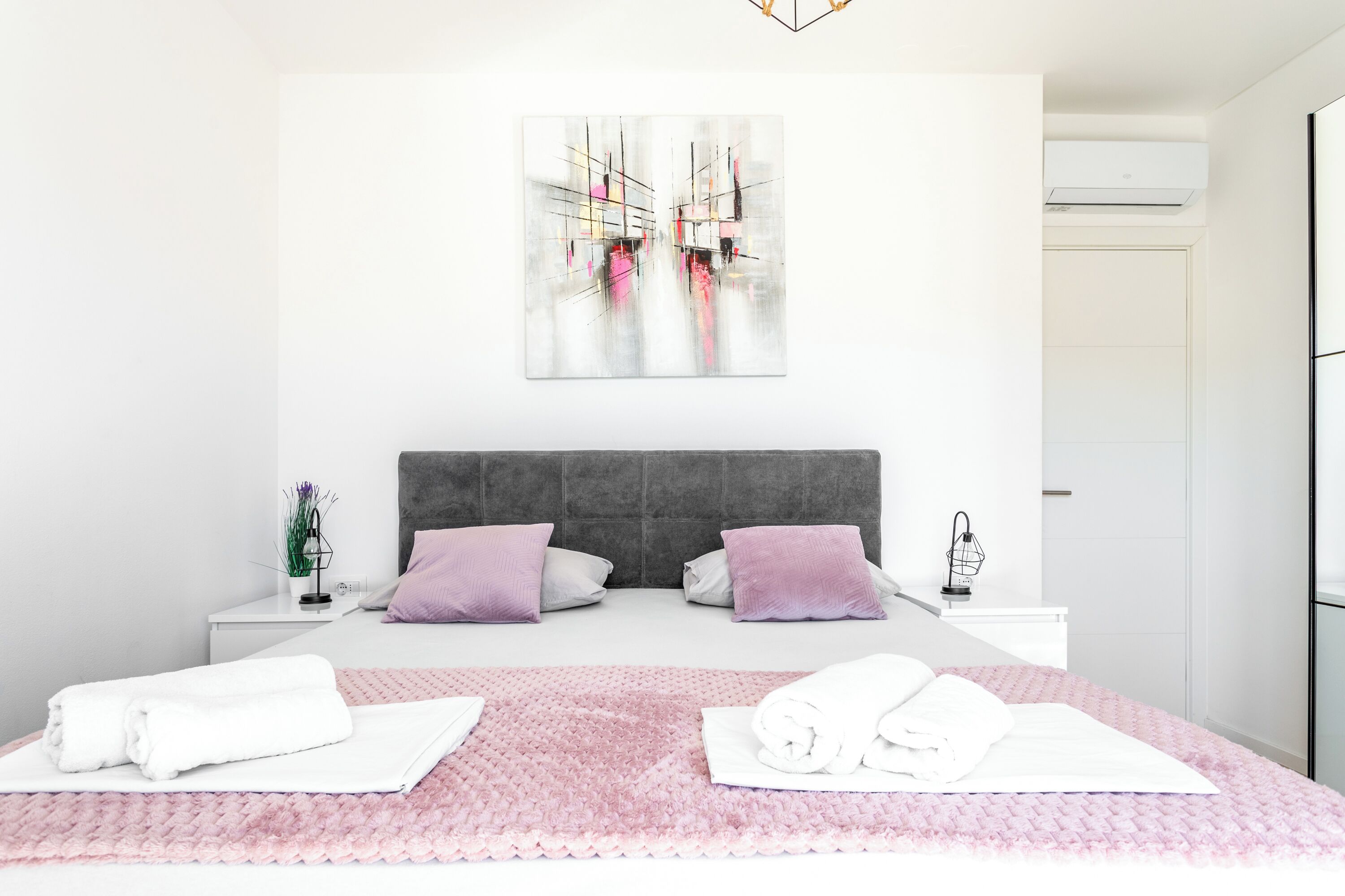 A modern hotel room with a large bed, two nightstands, a gray headboard, and a wall-mounted air conditioner.