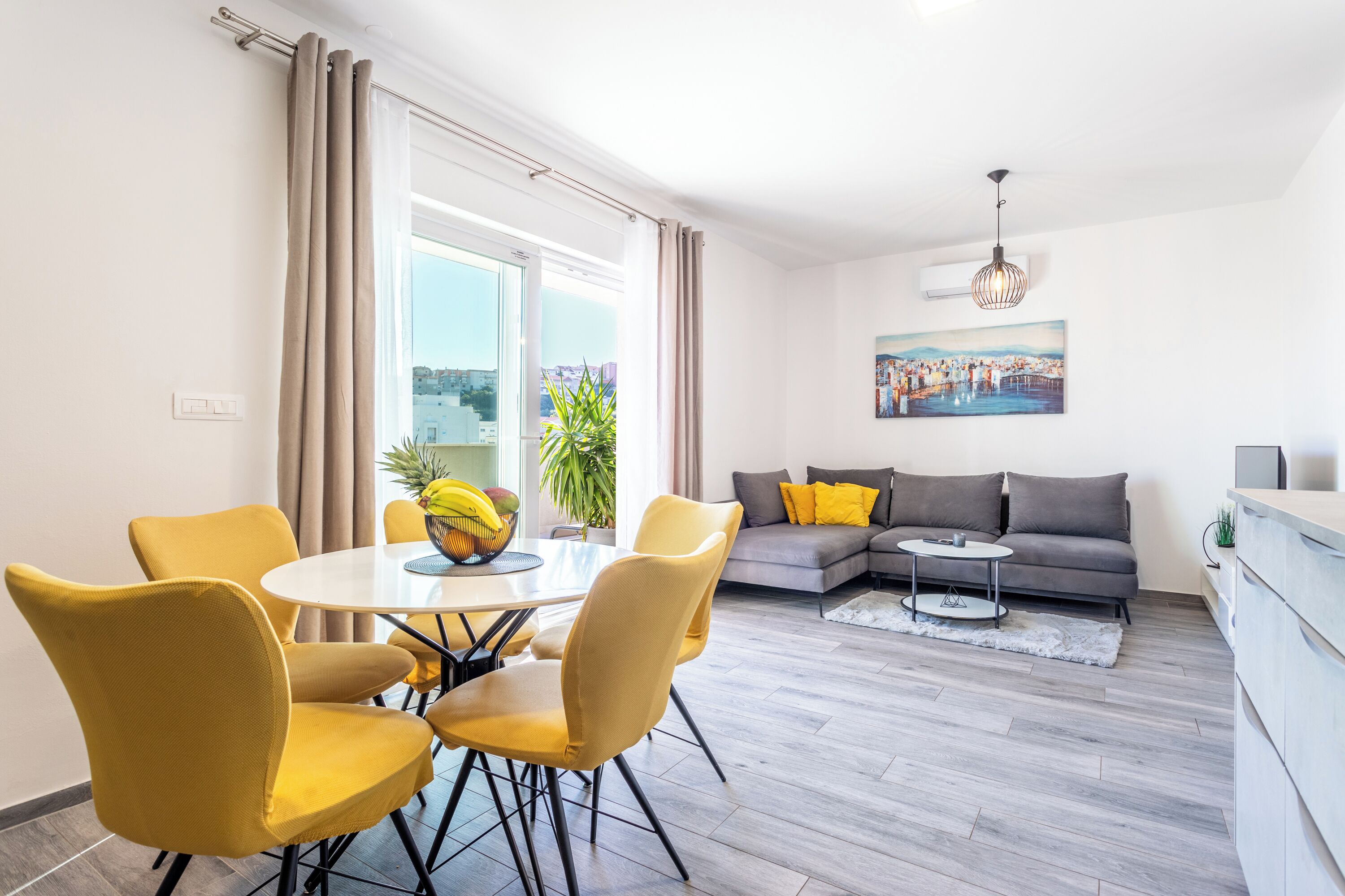 Una sala de estar moderna con zona de comedor, un sofá gris con cojines amarillos y un cuadro en la pared.