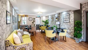 A hotel lobby with a yellow sofa, a round table, and chairs. There are framed pictures on the walls, a clock, and a sign that says "Good Vibes Only". - Eton House (Yeovil)