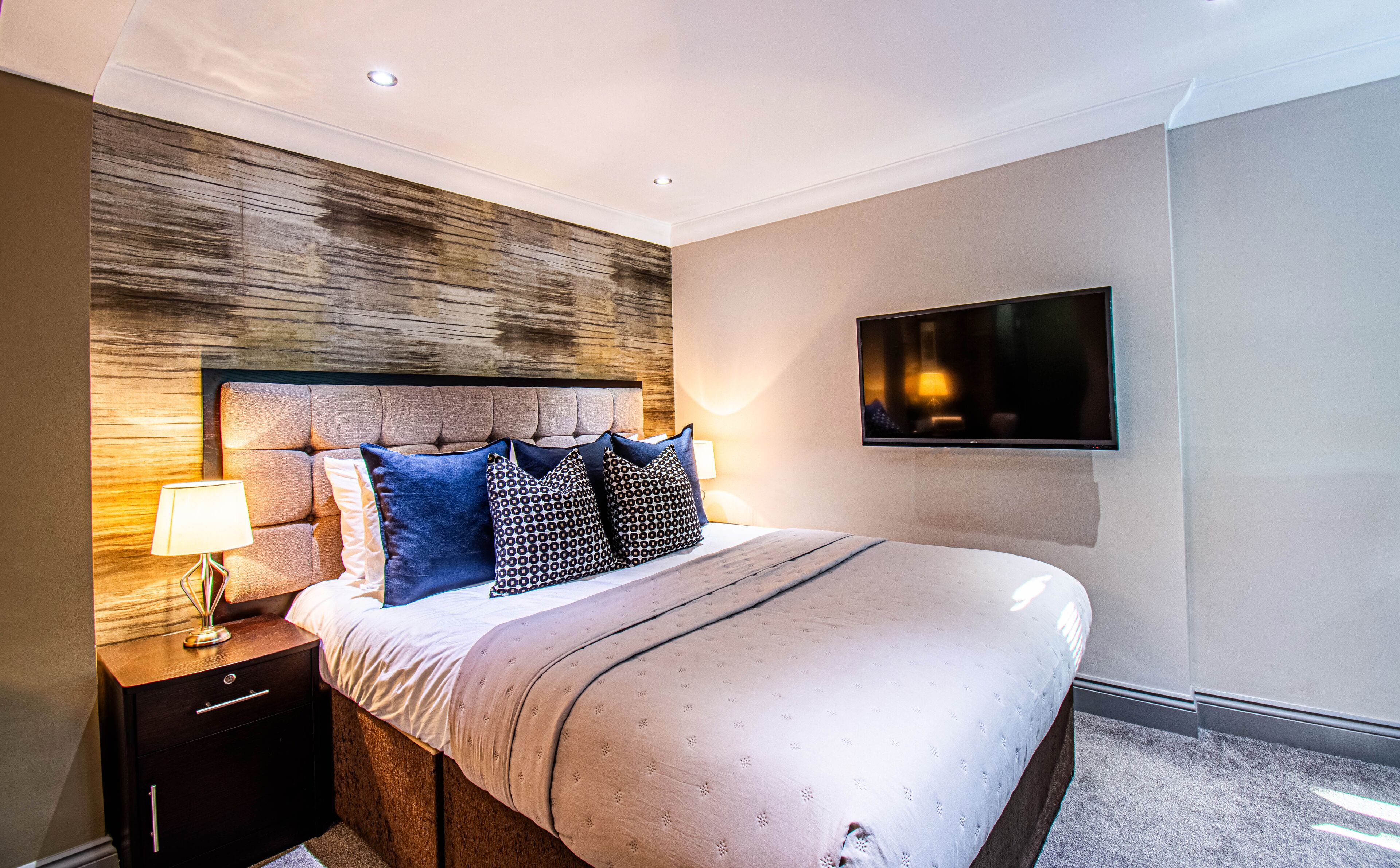 A modern hotel room with a large bed, a flat-screen TV mounted on the wall, a bedside table with a lamp, and a wooden accent wall.