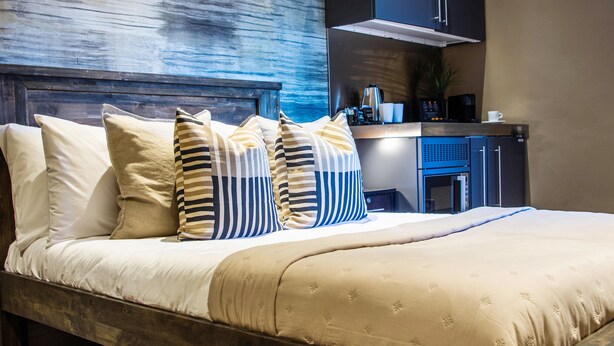 A neatly made bed with striped and solid pillows, a wooden headboard, and a modern kitchen area with a coffee machine and cabinets. - Eton House (Yeovil)